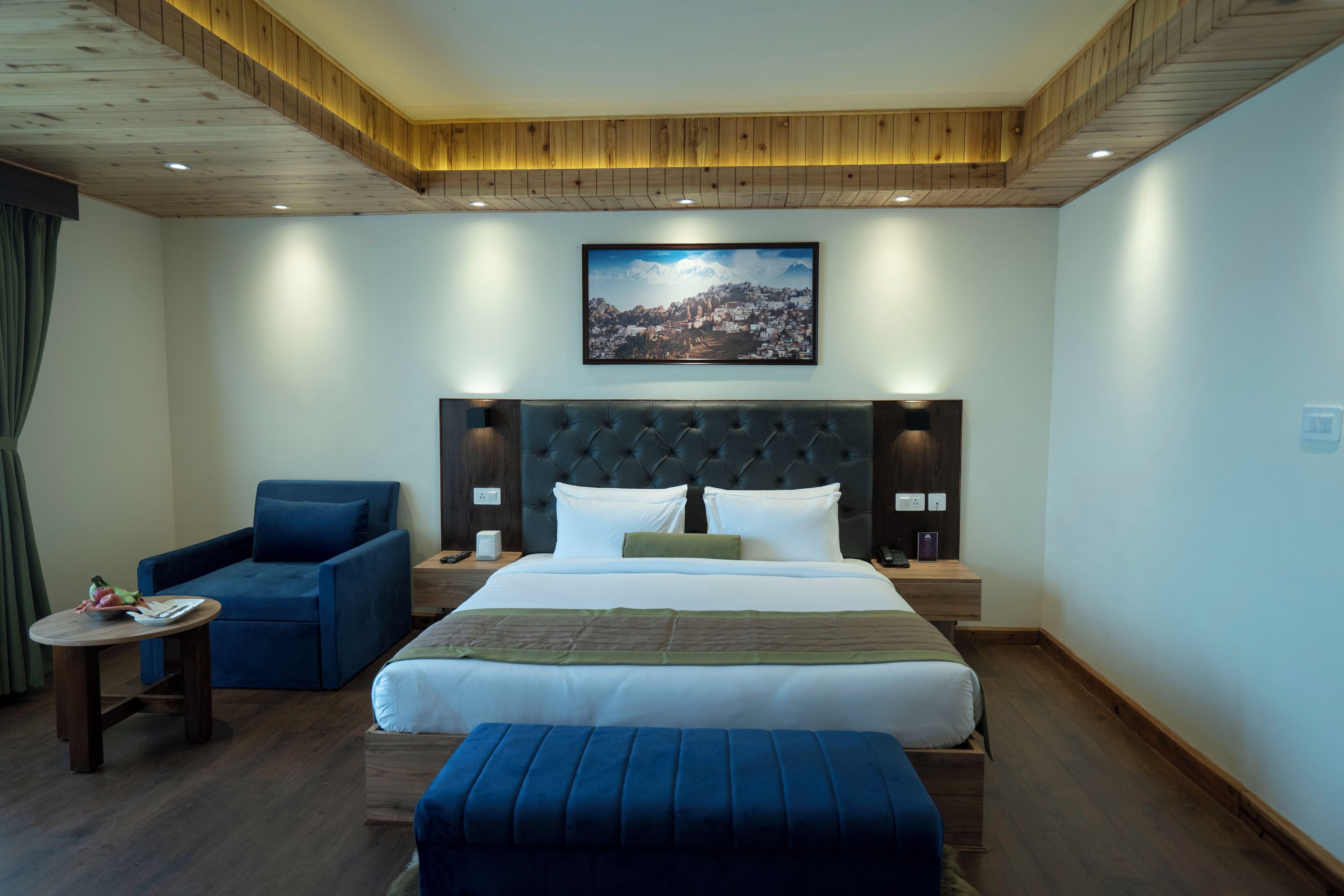 A hotel room with a large bed, a blue armchair, a small round table, and a framed picture on the wall.