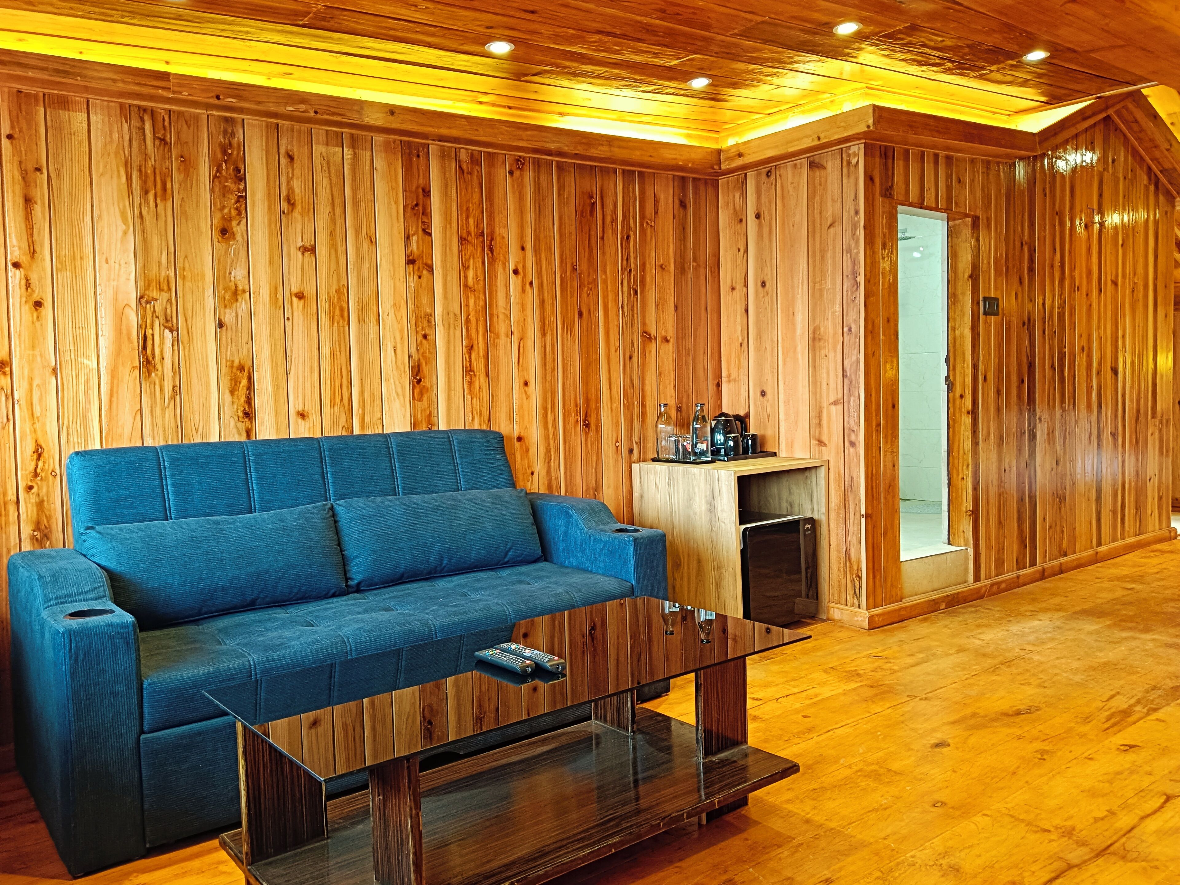 Une pièce aux murs lambrissés, avec un canapé bleu, une table basse en verre et un meuble en bois sur lequel sont disposés divers objets.
