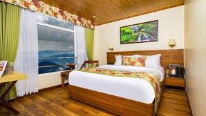Una habitación de hotel con una cama grande, cabecera de madera, mesita de noche con lámpara, una mesa con una silla y ventana con vistas a montañas.
