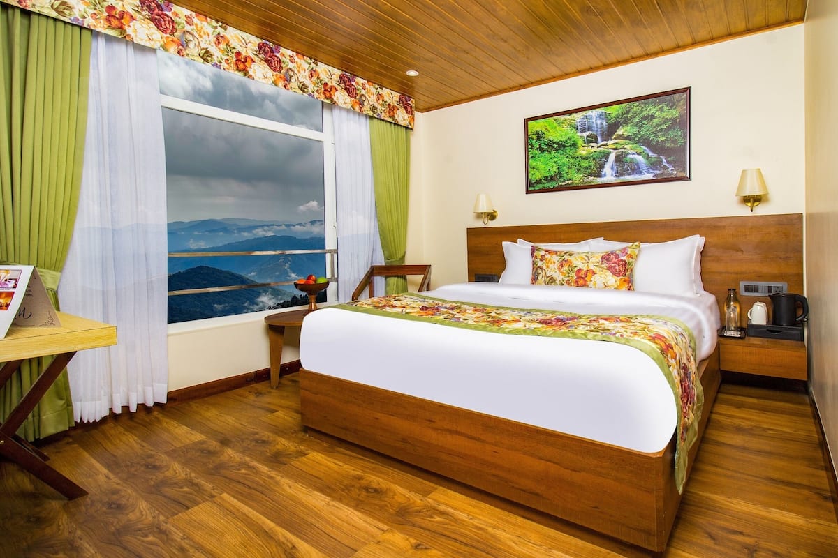 Una habitación de hotel con una cama grande, cabecera de madera, mesita de noche con lámpara, una mesa con una silla y ventana con vistas a montañas.
