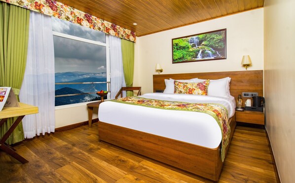A hotel room with a large bed, a wooden headboard, a nightstand with a lamp, a table with a chair, and a window with a view of mountains. - Valmere Lungta Boutique Hotel  (Darjeeling)