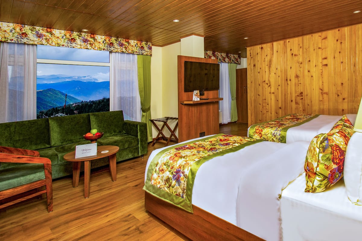 Habitación de hotel con techo de madera, un sofá verde, una cama con ropa de cama floral, una mesita de noche y una lámpara.