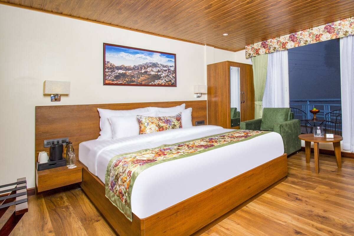 Habitación de hotel con una cama grande, cabecera de madera, mesita de noche con lámpara, una silla, una pequeña mesa y un balcón con vistas.