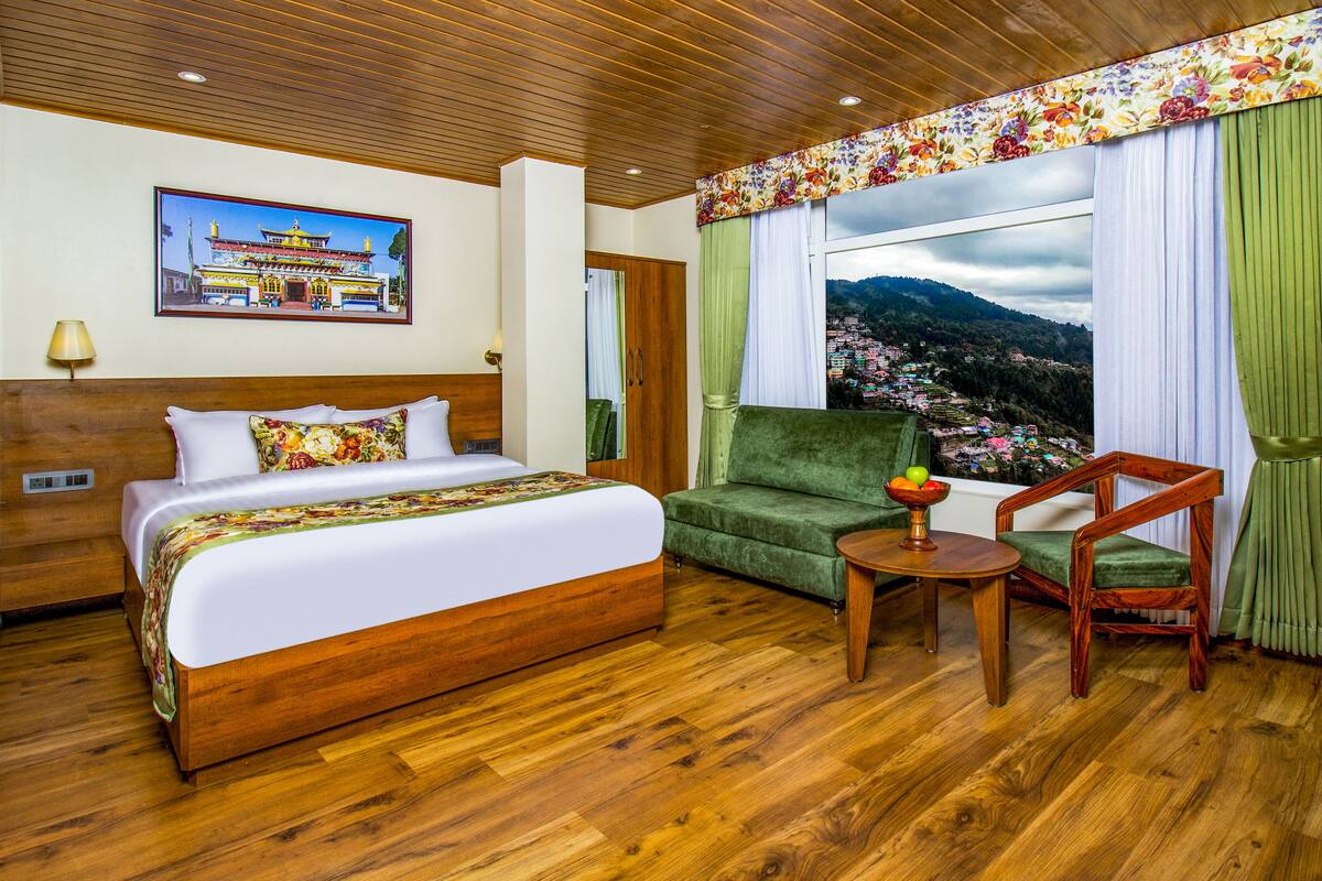Habitación de hotel con una cama de madera, un sillón verde, una mesita de noche de madera y un amplio ventanal con vistas a un paisaje montañoso.