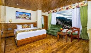 Quarto de hotel com cama de madeira, poltrona verde, mesa de cabeceira de madeira e uma janela ampla com vista para uma paisagem montanhosa.