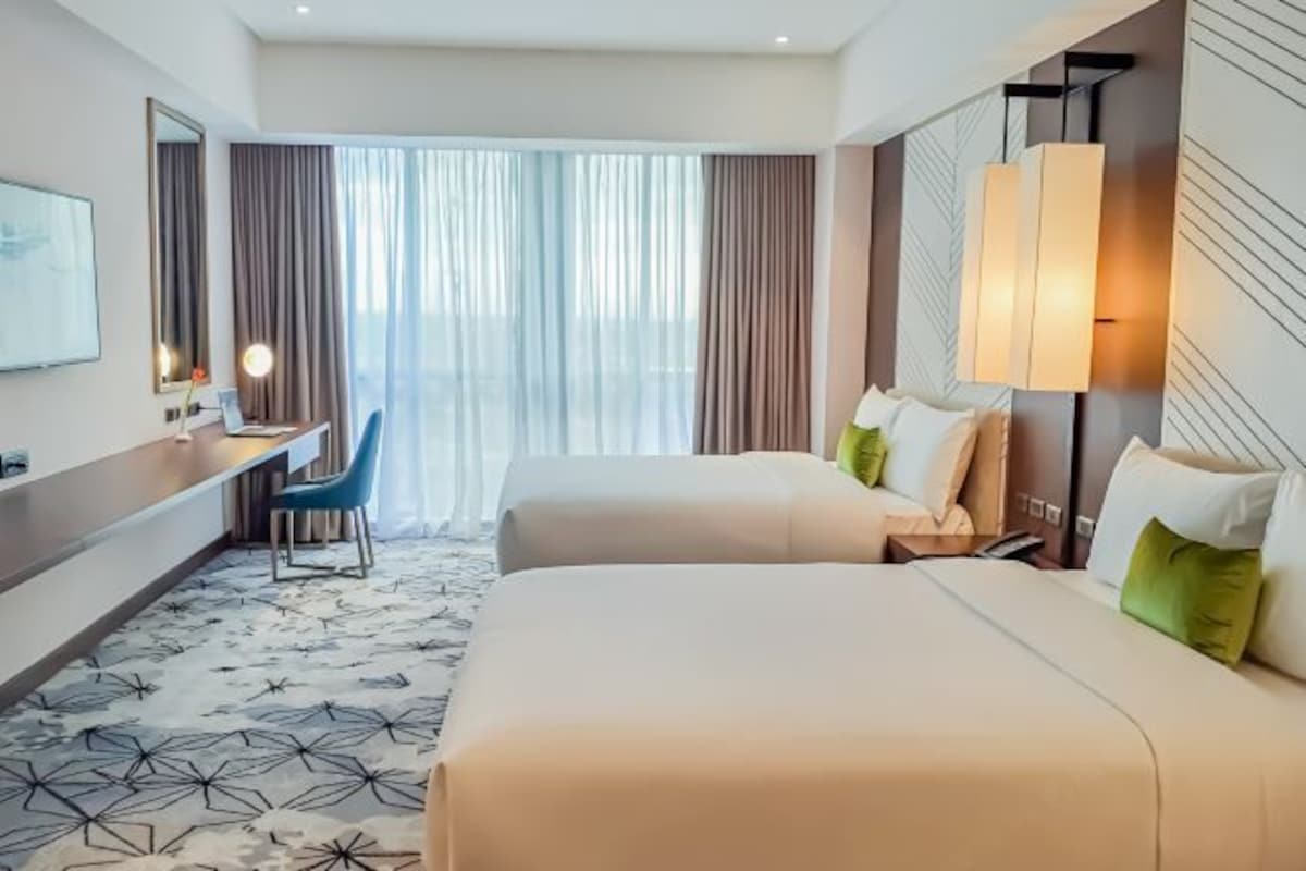 A hotel room with two beds, a desk, a chair, a large window with curtains, and a patterned carpet.