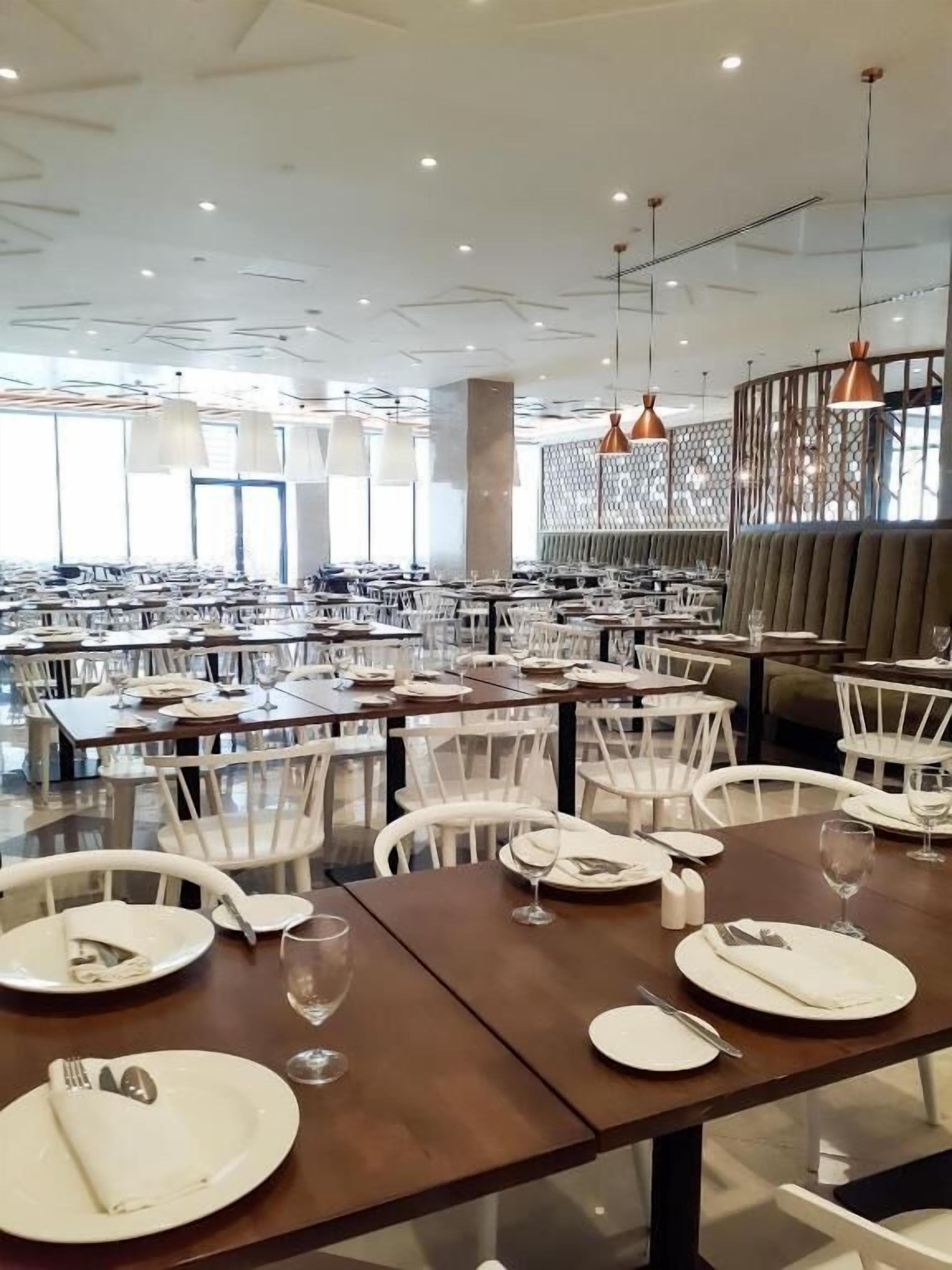 A spacious dining area with multiple tables and chairs, large windows, and pendant lighting.