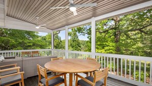 Eine überdachte Terrasse im Freien mit einem runden Holztisch und Stühlen, umgeben von einem weißen Geländer und Blick ins Grüne.