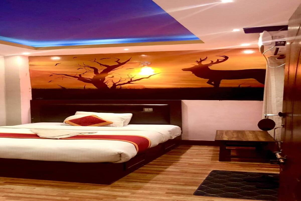 Une chambre d’hôtel moderne avec un grand lit, une tête de lit en bois, une fresque représentant un arbre et des cerfs peinte au plafond, et une petite table placée près du lit.
