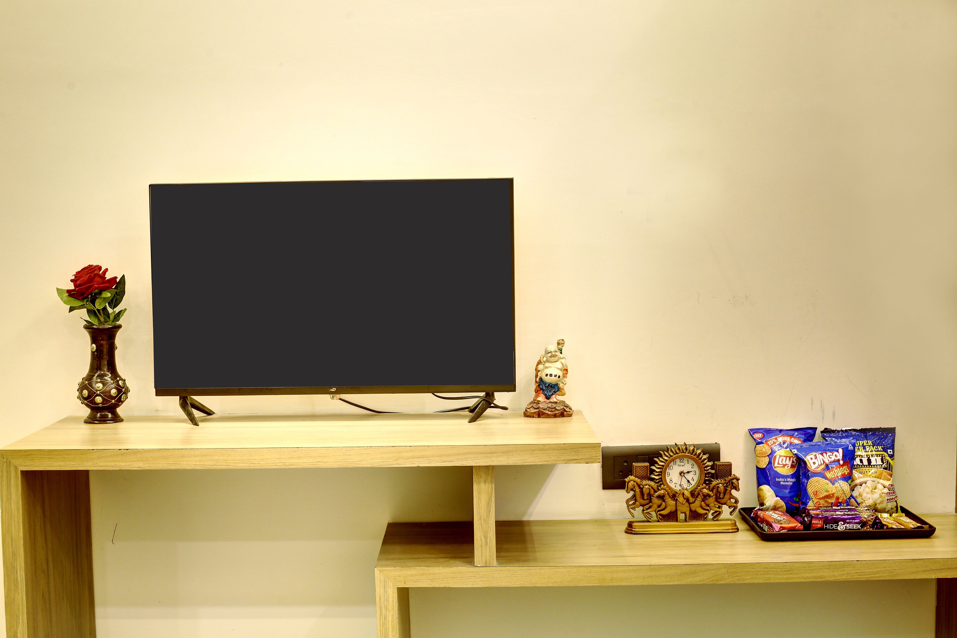 A wooden desk with a television, a vase with a red rose, a clock, and snacks.
