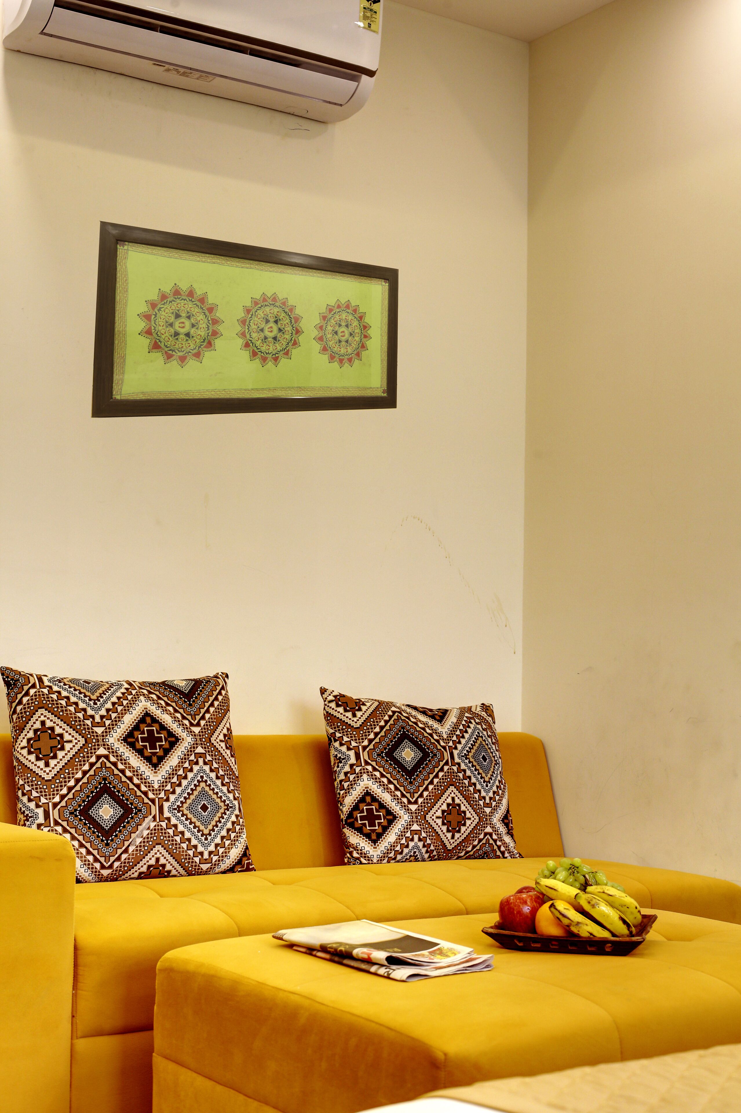 Un sofá amarillo con almohadones estampados, una obra de arte enmarcada encima y un frutero sobre el sofá.