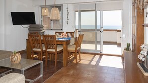 A dining area with a wooden table and chairs, a TV mounted on the wall, and a view of the sea through a sliding glass door.