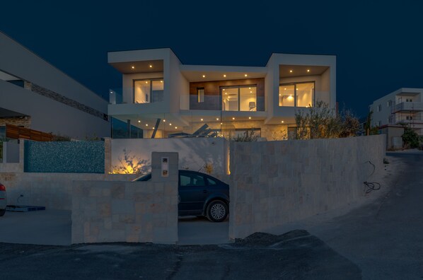 Una casa moderna de dos pisos con un coche estacionado en la entrada, rodeada por un muro de piedra.