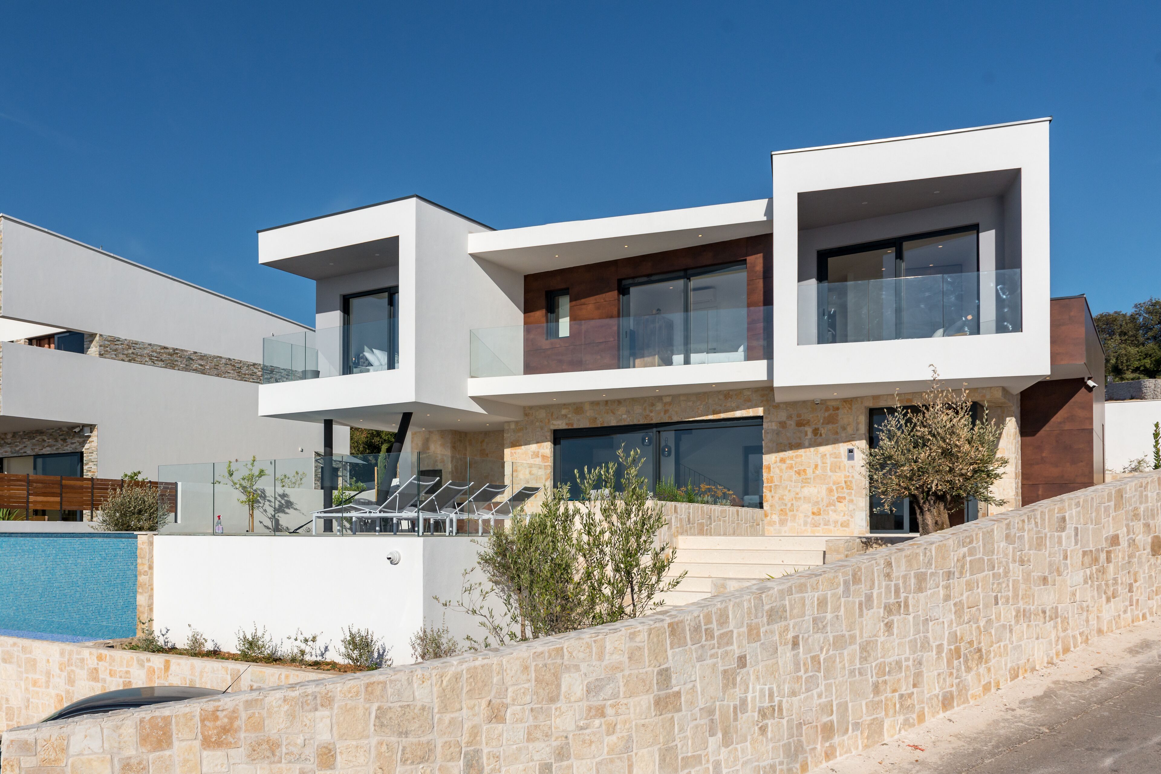 A modern two-story house with a stone retaining wall, a swimming pool, and a balcony with sun loungers.