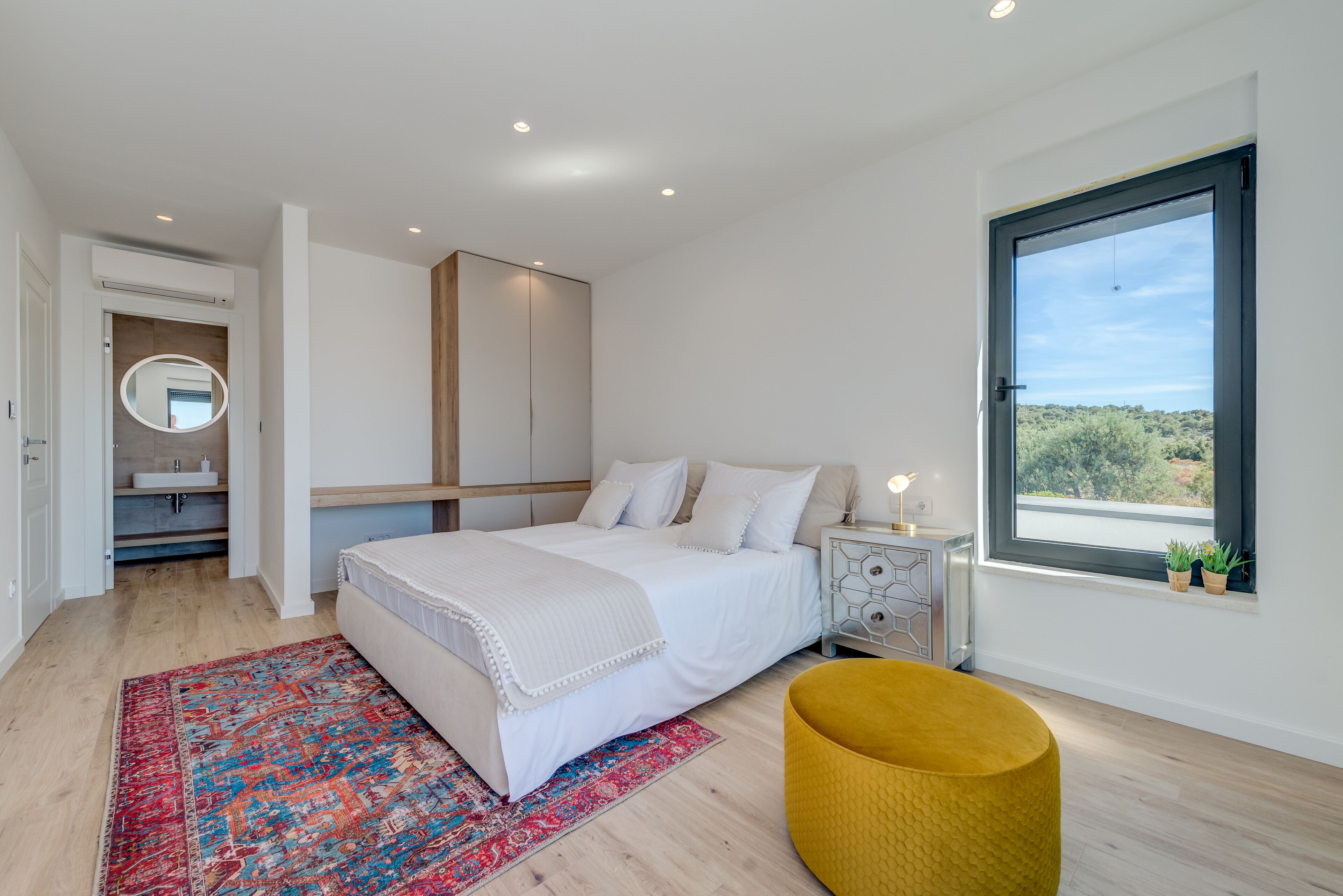 Une chambre moderne avec un grand lit, un miroir rond, une table de chevet et une fenêtre donnant sur un beau paysage.
