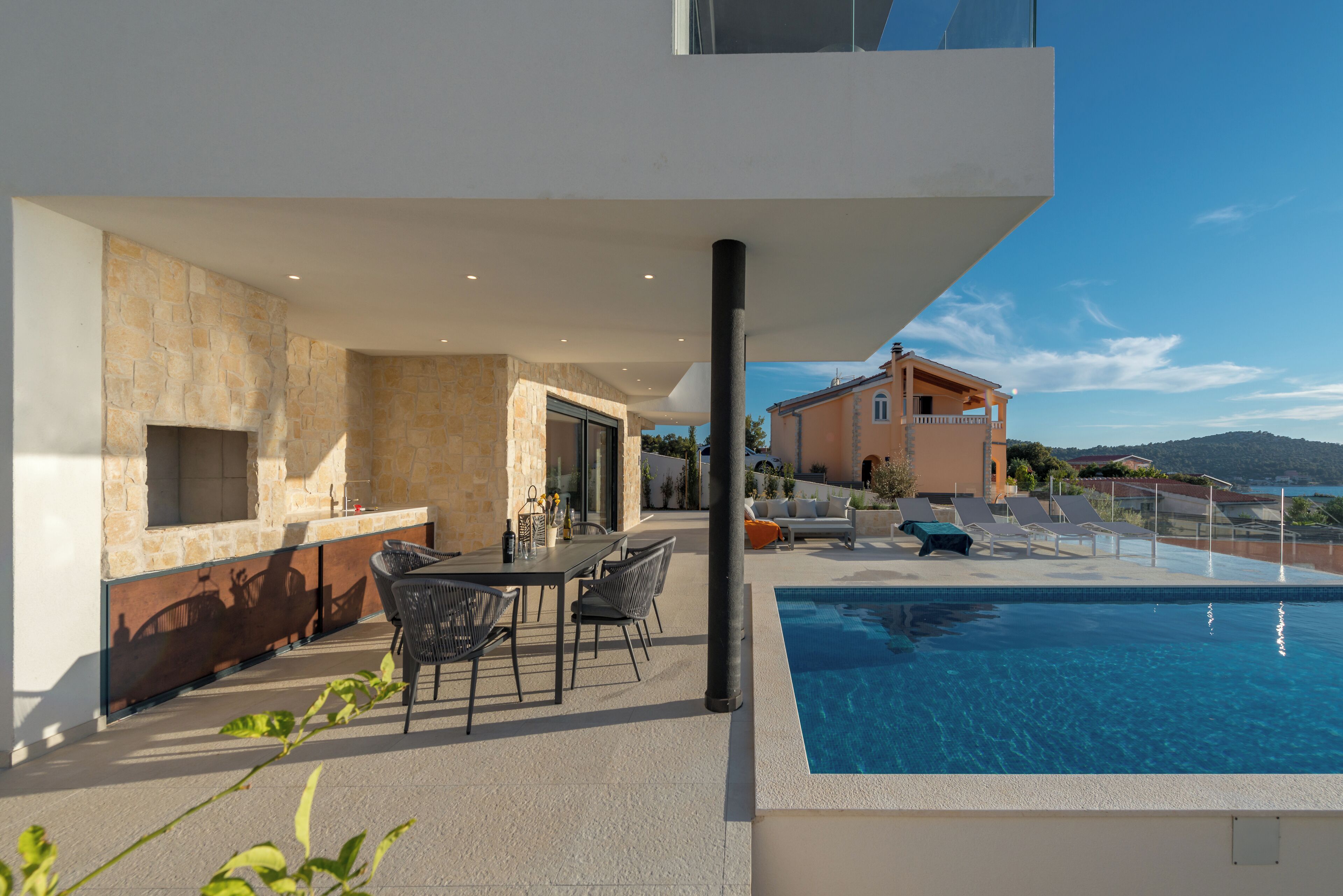 A modern outdoor pool area with a stone wall, a dining table with chairs, and a view of a building in the background.