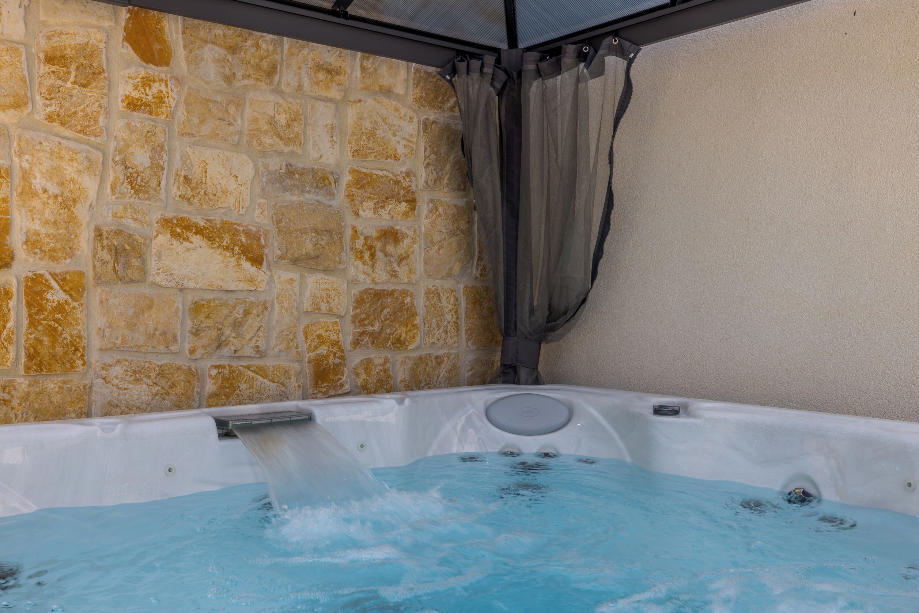 A hot tub with a stone wall backdrop and a grey curtain.