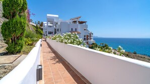 A coastal residential building with a terracotta-tiled pathway, white walls, and a view of the sea.