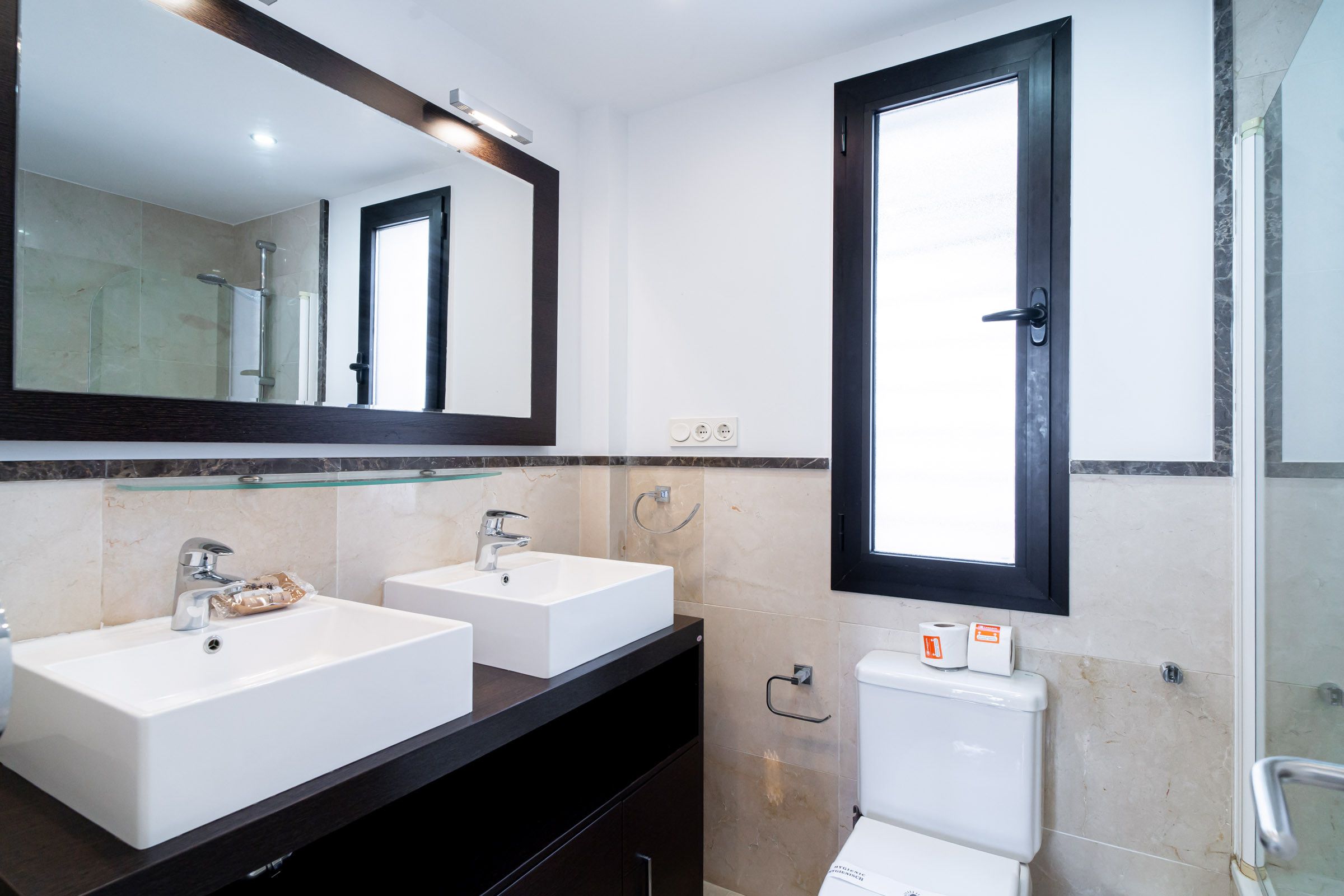 A bathroom with two sinks, a mirror, a toilet, and a window.