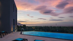 A modern pool area with a clear view of the ocean at sunset, a seating area with lounge chairs, and a well-lit exterior wall. - Grand Luxe Villa - Sea views, hot tub, private pool & modern design (Chania)