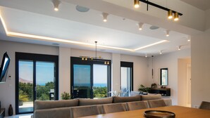 A modern living room with a large wooden dining table, a sofa, and a view of the outdoors through large sliding doors. - Grand Luxe Villa - Sea views, hot tub, private pool & modern design (Chania)
