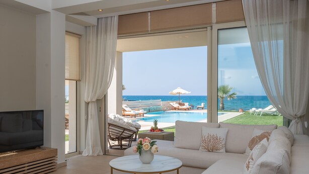 Ein modernes Wohnzimmer mit großem Fernseher, einer Couch, zwei runden Tischen und Blick auf Pool und Meer.