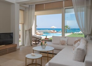 A modern living room with a large TV, a sofa, two round tables, and a view of a pool and ocean. - Unique beachfront location, heated pool with built-in hydromassage, large garden (Heraklion)