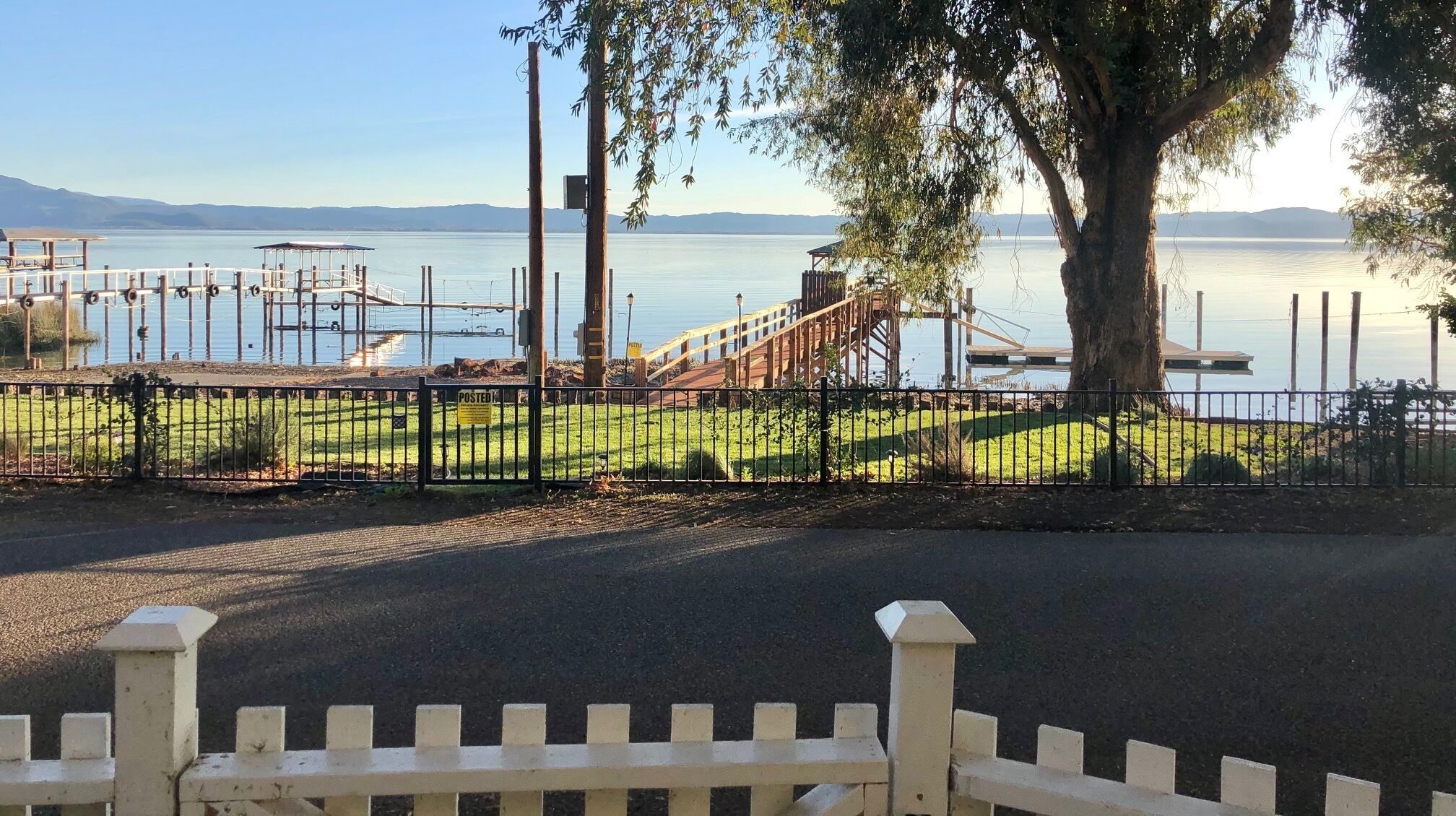 Ein von einer Hecke umgebener Parkbereich mit Spielplatz, einem großen Baum und Blick auf ein Gewässer.