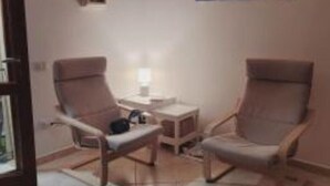 Ein Zimmer mit zwei Sesseln, einem kleinen Tisch mit einer Lampe, einem Gemälde an der Wand und einem Holzkabinett.