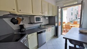A compact kitchen with white cabinets, a microwave, and a dishwasher. There is a dining area with a table and chairs visible through an open door. - Appartement 2 Avec Emplacement Idéal et Clim (Collioure)