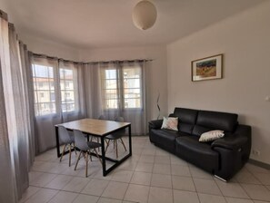 Uma sala de estar com um sofá de couro preto, uma mesa de jantar de madeira com cadeiras e janelas amplas com cortinas leves.