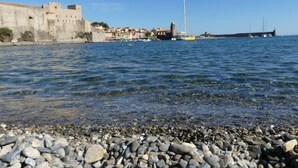 A coastal scene with a rocky beach, a historic stone wall, and a sailboat in the water. - Appartement 2 Avec Emplacement Idéal et Clim (Collioure)