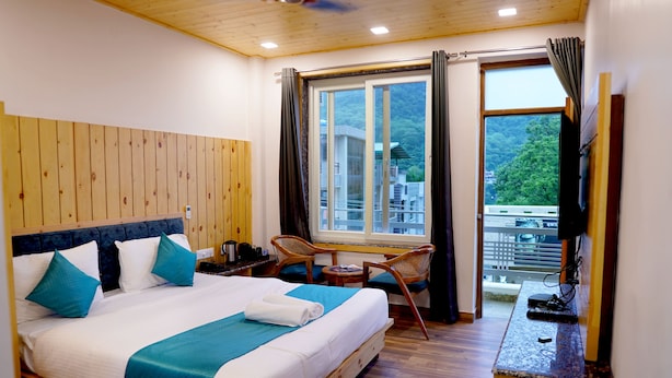 Una habitación de hotel con cabecera de madera, una cama grande con ropa de cama azul y blanca, una pequeña mesa con sillas, un televisor y un balcón con vistas a edificios y árboles.