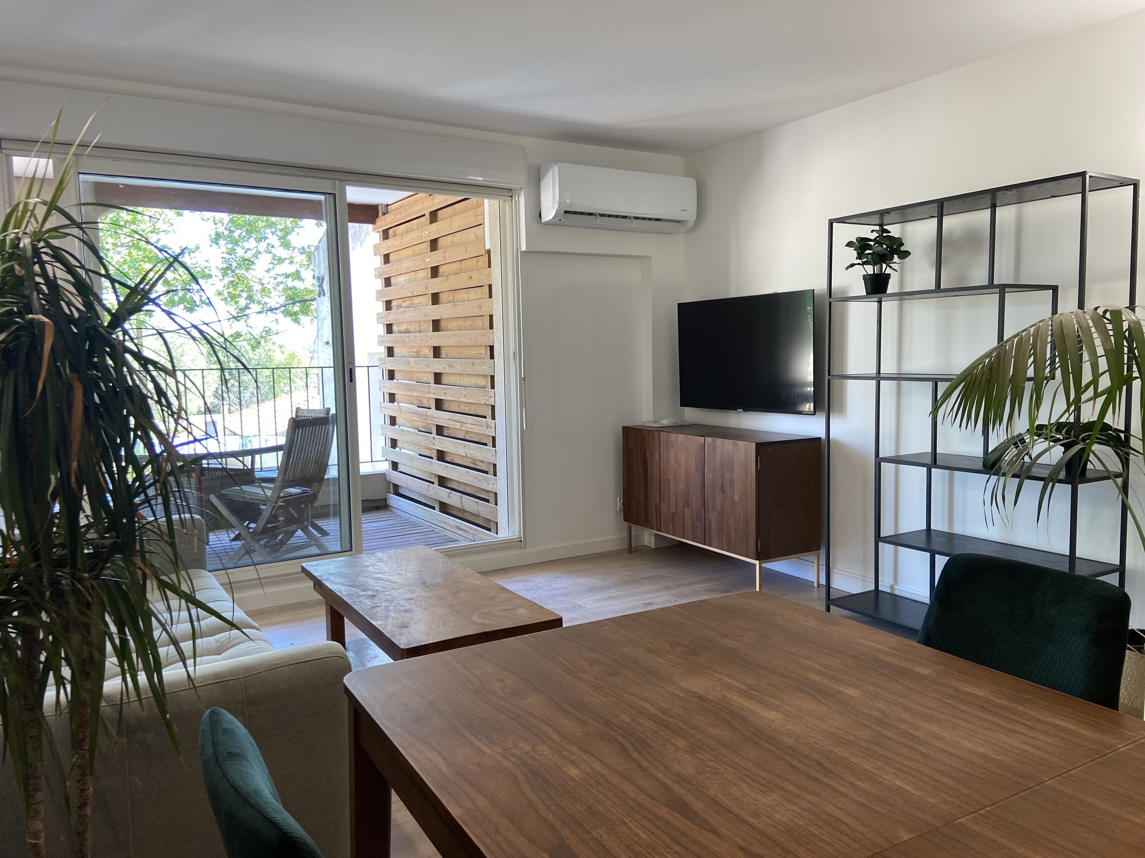 Un soggiorno moderno con un tavolo da pranzo in legno, una televisione su un mobiletto in legno e un balcone con ringhiera in legno.