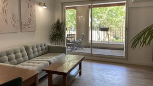 A living room with a sofa, a wooden table, and a sliding glass door leading to an outdoor area with a chair and a plant. - T2 Bord du Canal du Midi Piscine (Homps)