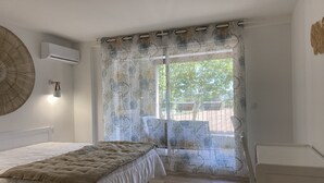 A bedroom with a bed, a bedside lamp, a window with patterned curtains, and a view of the outdoors. - T2 Bord du Canal du Midi Piscine (Homps)