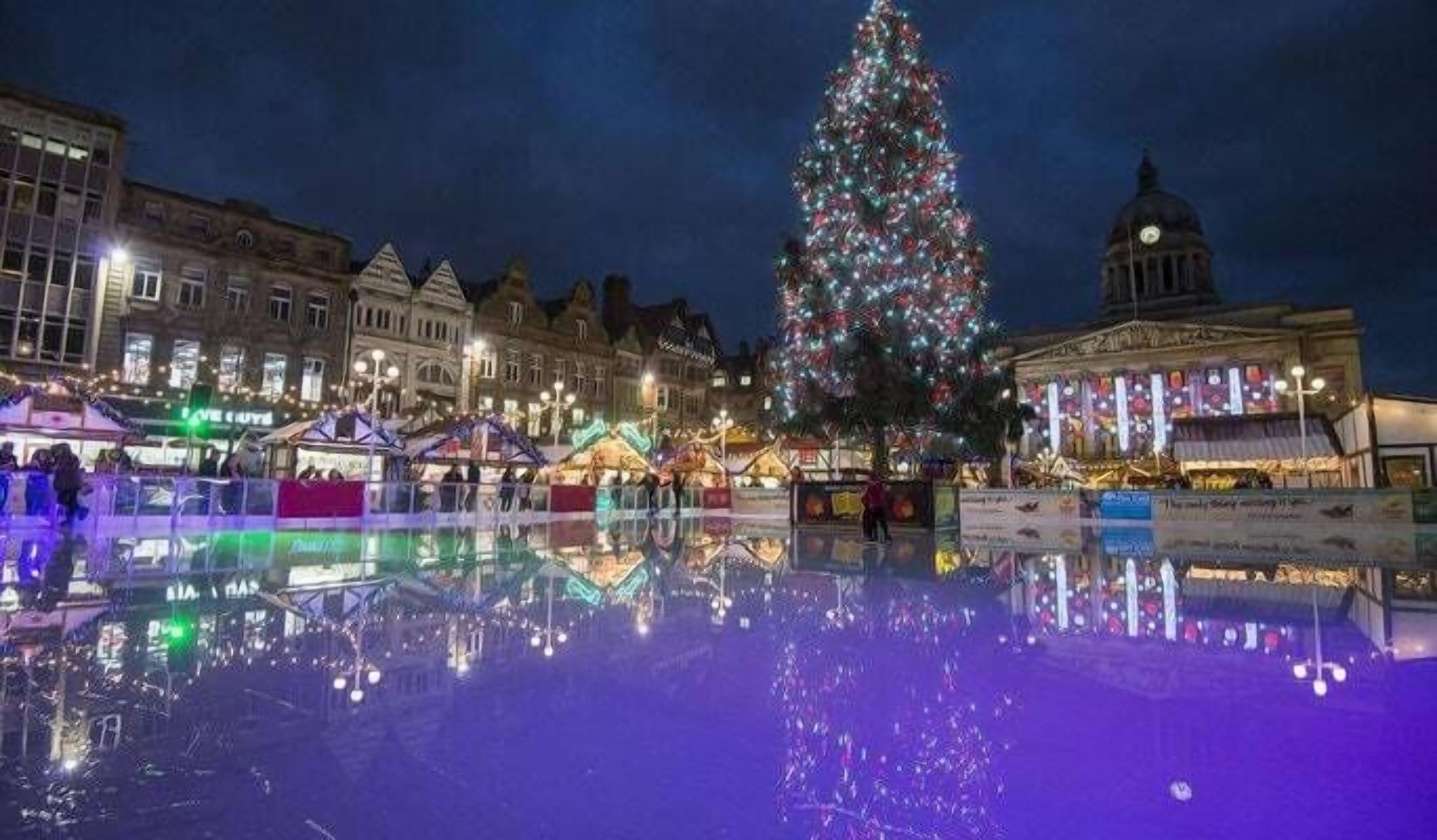 Une place de ville illuminée la nuit, avec un grand sapin de Noël, des bâtiments éclairés et une surface d’eau qui reflète la lumière.