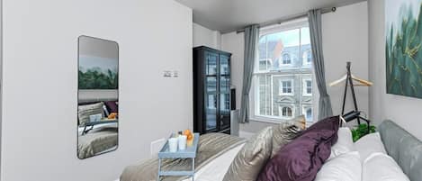 Ein Schlafzimmer mit einem Bett, einem Nachttisch mit Tablett, einem Spiegel, einem Schrank und einem Fenster mit Vorhängen.