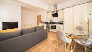 A modern living room with a sofa, a glass dining table, and a kitchen area with cabinets and appliances. - Wow! 1bed Stunning Apt in Victorian Mansion (Nottingham)
