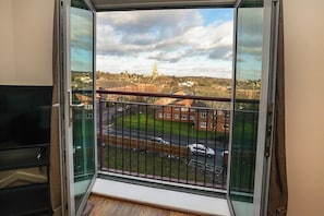 A room with a large window overlooking a street and a church, a balcony with a metal railing, and a television on the left.