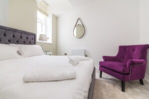 Un lit bien fait, avec du linge de lit blanc, un miroir rond accroché au mur et un fauteuil violet.