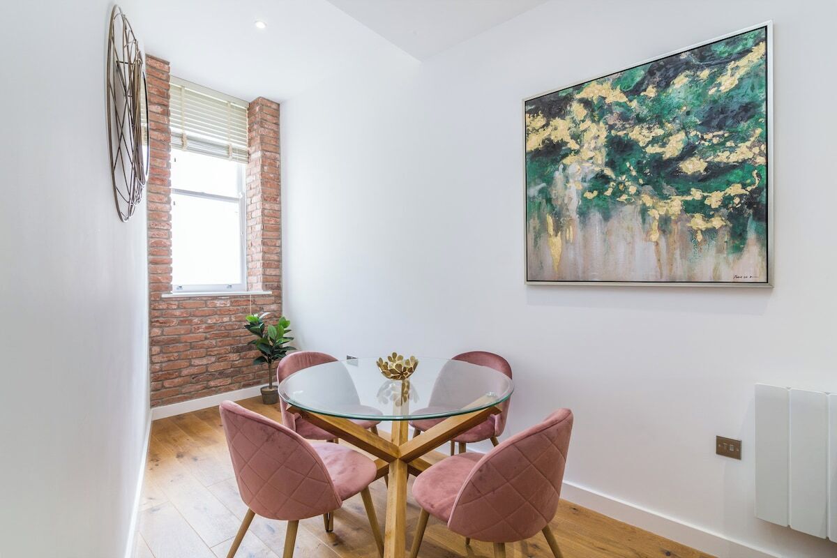 A dining area with a round glass table, pink upholstered chairs, a brick wall, and a large abstract painting.