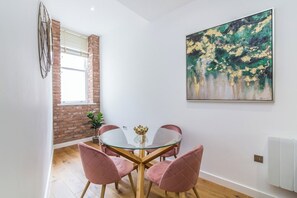 A dining area with a round glass table, pink upholstered chairs, a brick wall, and a large abstract painting. - Lovely 1 Bedroom Apartment at City Centre (Nottingham)