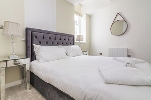 A neatly made bed with white linens, a grey upholstered headboard, and a bedside table with a lamp. - Lovely 1 Bedroom Apartment at City Centre (Nottingham)