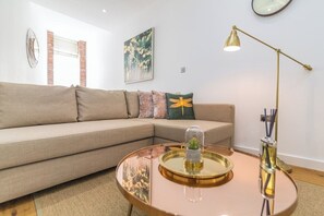 Un salon moderne avec un canapé d’angle beige, une table basse ronde avec un plateau en verre, un lampadaire et une horloge murale.