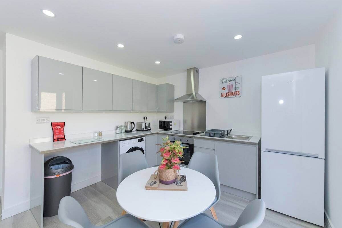 A modern kitchen with white cabinets, a stainless steel range hood, a refrigerator, a microwave, and a coffee maker. There is a small dining table with chairs and a trash bin.