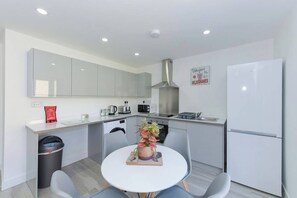 Eine moderne Küche mit weißen Schränken, einer Edelstahl-Dunstabzugshaube, einem Kühlschrank, einer Mikrowelle und einer Kaffeemaschine. Es gibt einen kleinen Esstisch mit Stühlen und einen Mülleimer.