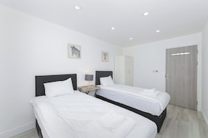 A hotel room with two single beds, a nightstand with a lamp, a wardrobe, and a door. - Gedling 2-BR Apartment w/ Easy Parking (Gedling)