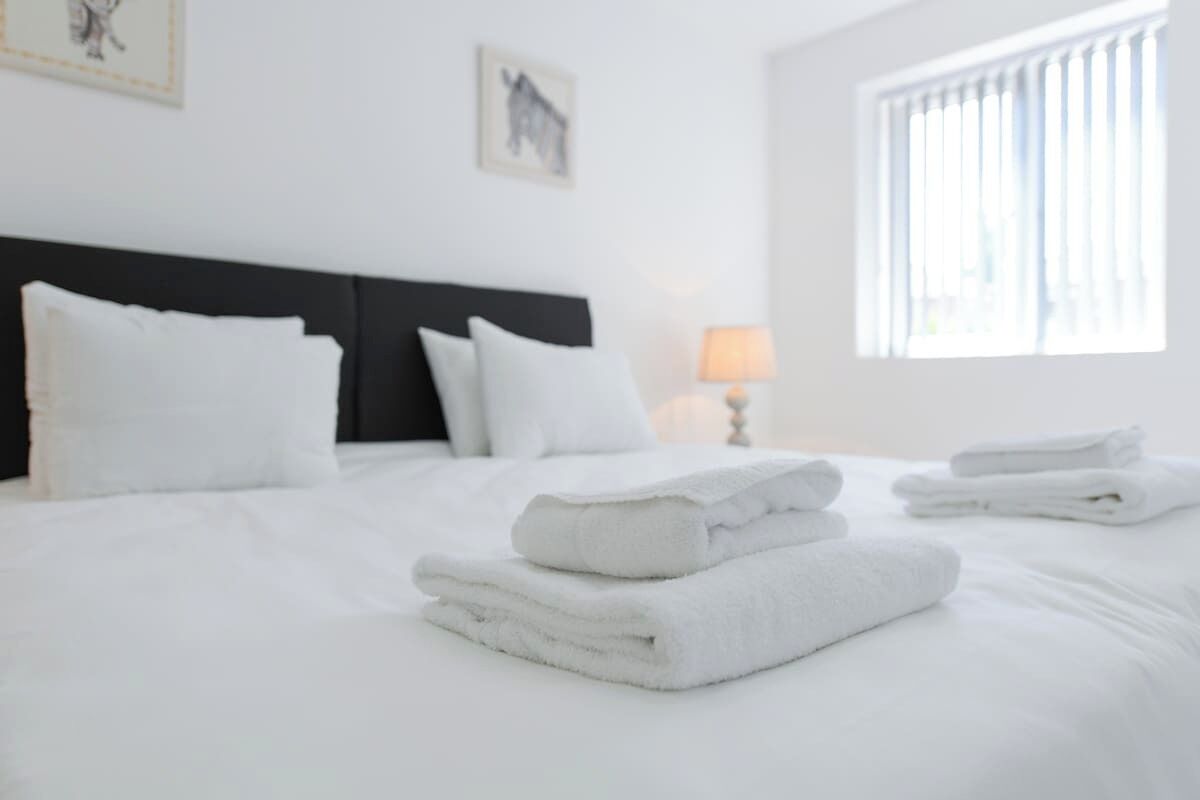 A neatly made bed with white linens and towels, a black headboard, and a window with blinds.