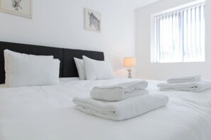 A neatly made bed with white linens and towels, a black headboard, and a window with blinds. - 2-BR Apartment in Gedling, Close to City (Gedling)